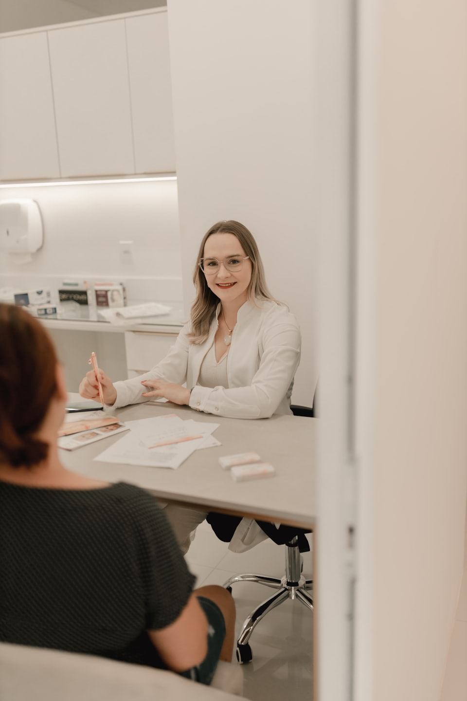 Woman in lab coat smiles at desk with pencil.